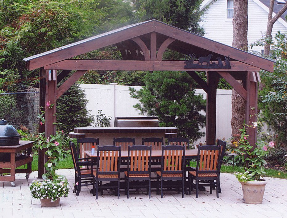 Açık havada, ahşap bir gazebo altında yemek masası ve sandalyeleri bulunan bir barbekü alanı. Arka planda yeşillikler ve beyaz bir çit görülmektedir.