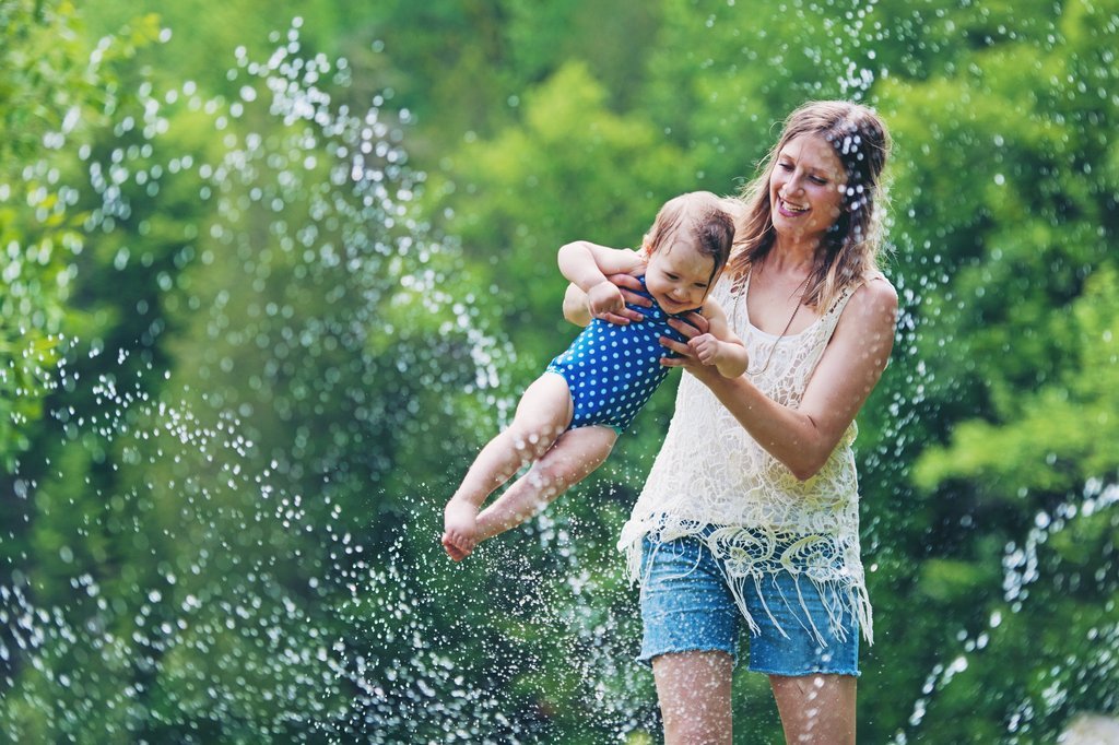 Anne, bahçede bebeğiyle birlikte suyla oynarken neşeli bir anı yakalıyor. Bebek mavi puantiyeli mayosuyla annesinin kollarında gülümsüyor.