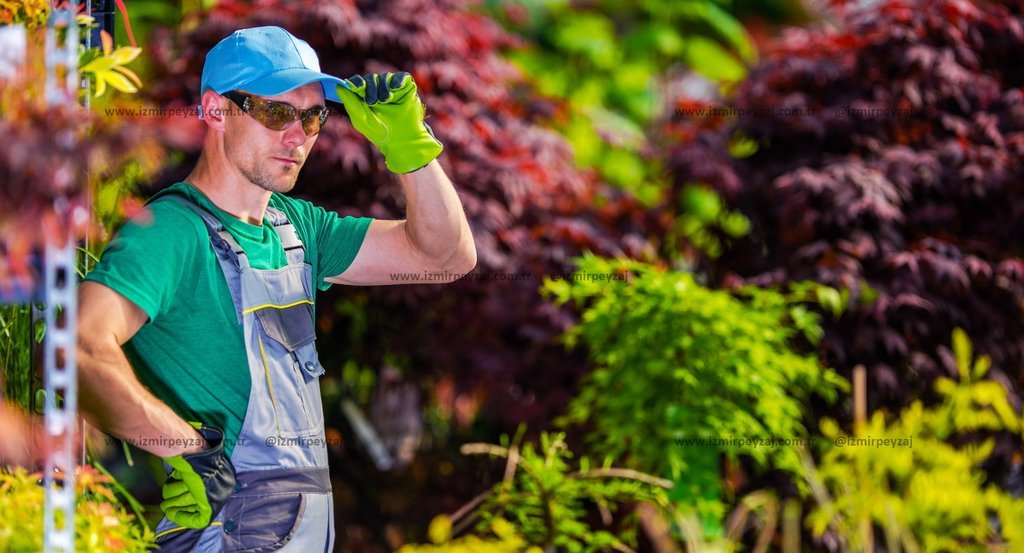 Bahçıvan kıyafetleri giymiş bir adam, bahçede çalışırken güneş gözlüğü ve şapka takıyor. Arka planda Bahçıvan kıyafetleri giymiş bir adam, bahçede çalışırken güneş gözlüğü ve şapka takıyor. Arka planda çeşitli bitki türleri ve renkli yapraklar görülüy