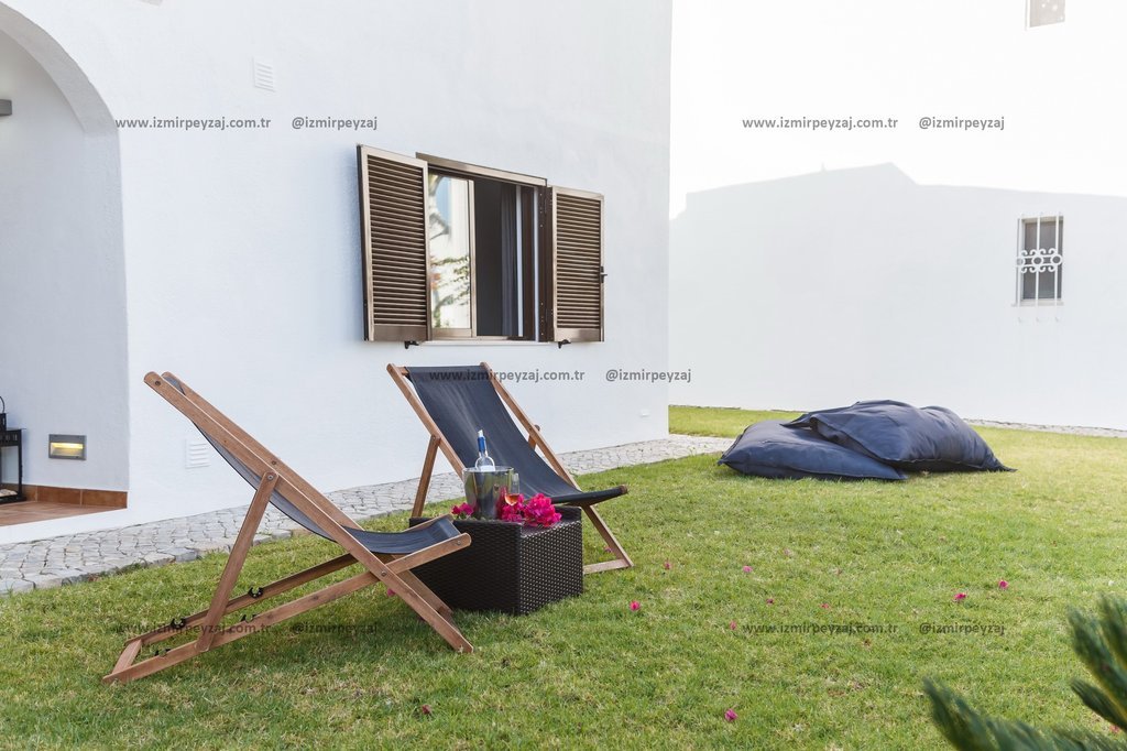 Bu görselde, bahçede yer alan şezlonglar, şarap ve çiçeklerle süslenmiş bir masa ile huzurlu bir atm Bu görselde, bahçede yer alan şezlonglar, şarap ve çiçeklerle süslenmiş bir masa ile huzurlu bir atmosfer sunuluyor. Beyaz duvarlar ve yeşil çimlerle