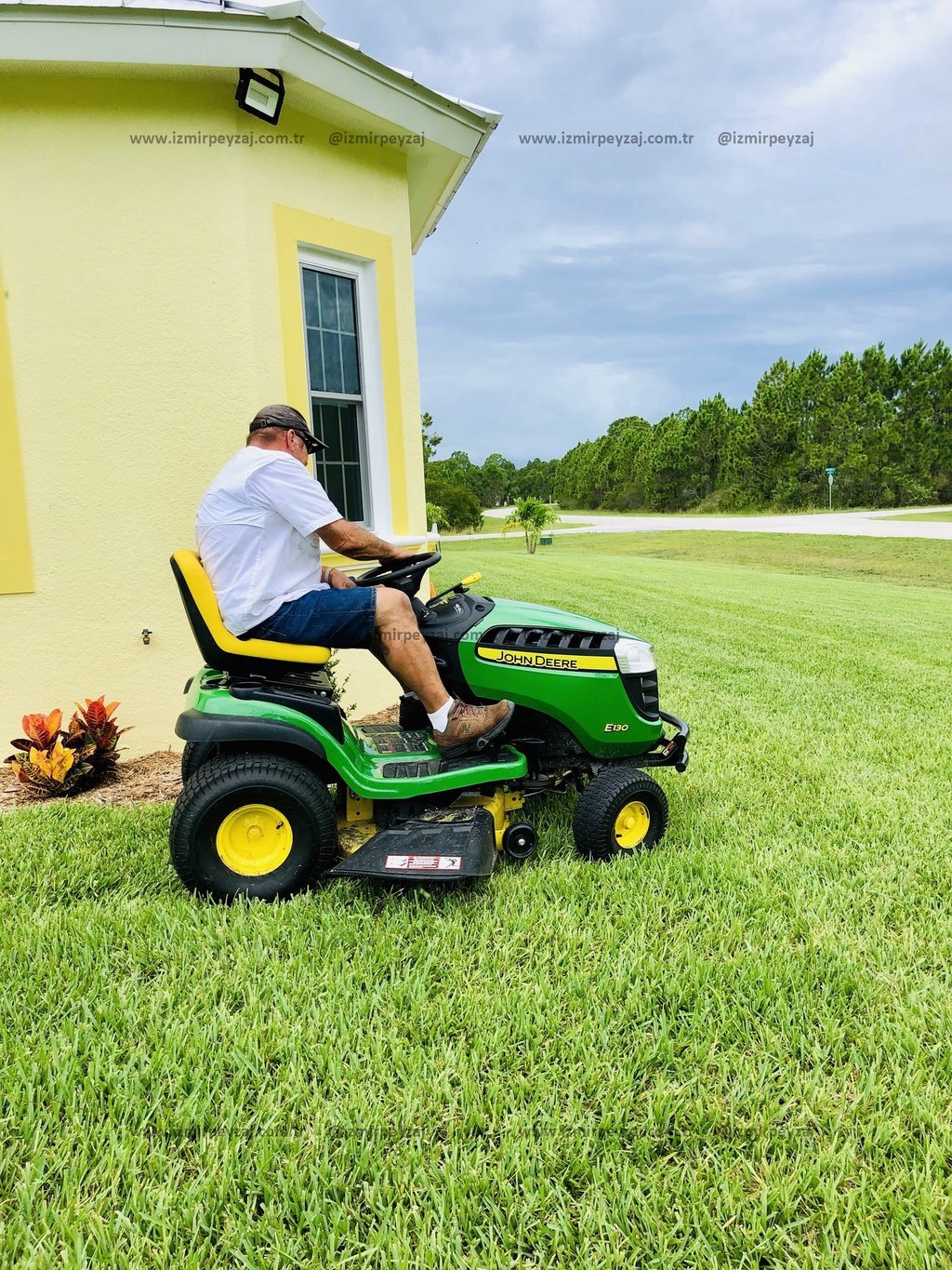 Bahçesinde çim biçme makinesi kullanan bir adamın görüntüsü. Yeşil çimler ve ağaçlarla dolu bir peyzajda, sarı bir evin yanında çim biçme işlemi yapıl