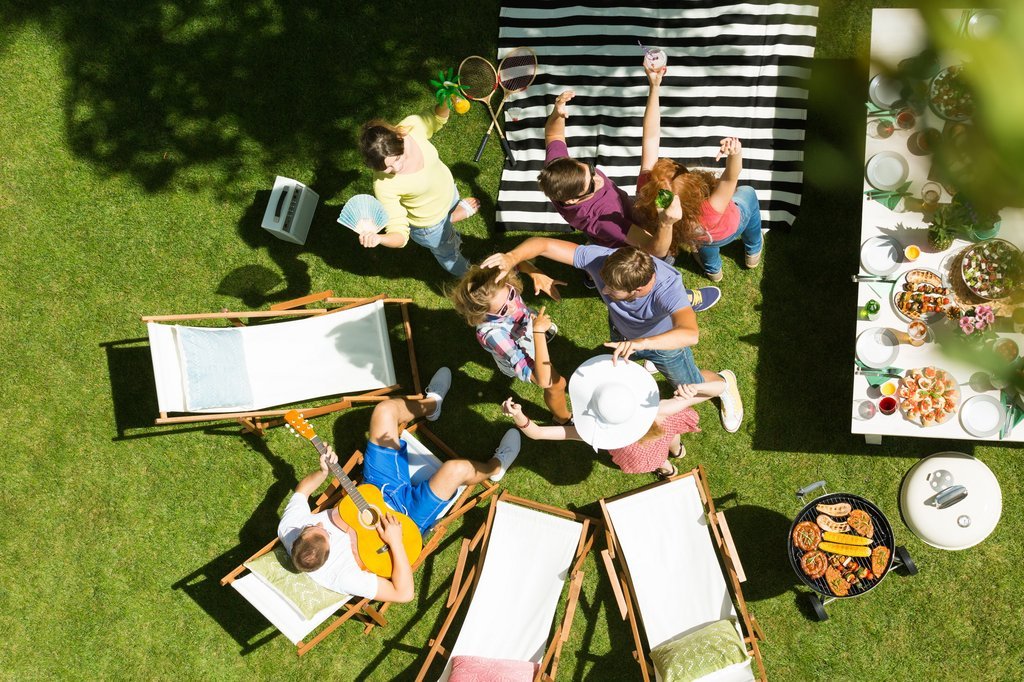 Yeşil bir bahçede, güneşli bir günde piknik yapan bir grup insanın yukarıdan çekilmiş fotoğrafı. İnsanlar neşeyle sohbet ediyor, müzik dinliyor ve yem
