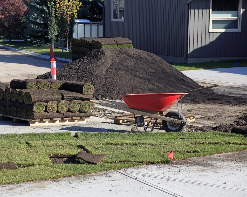 Resimde, yeni çim serilmesi için hazırlanan bir alan ve gerekli malzemeler görülmektedir. Kırmızı bir el arabası, toprak yığını ve rulo çimler dikkat 