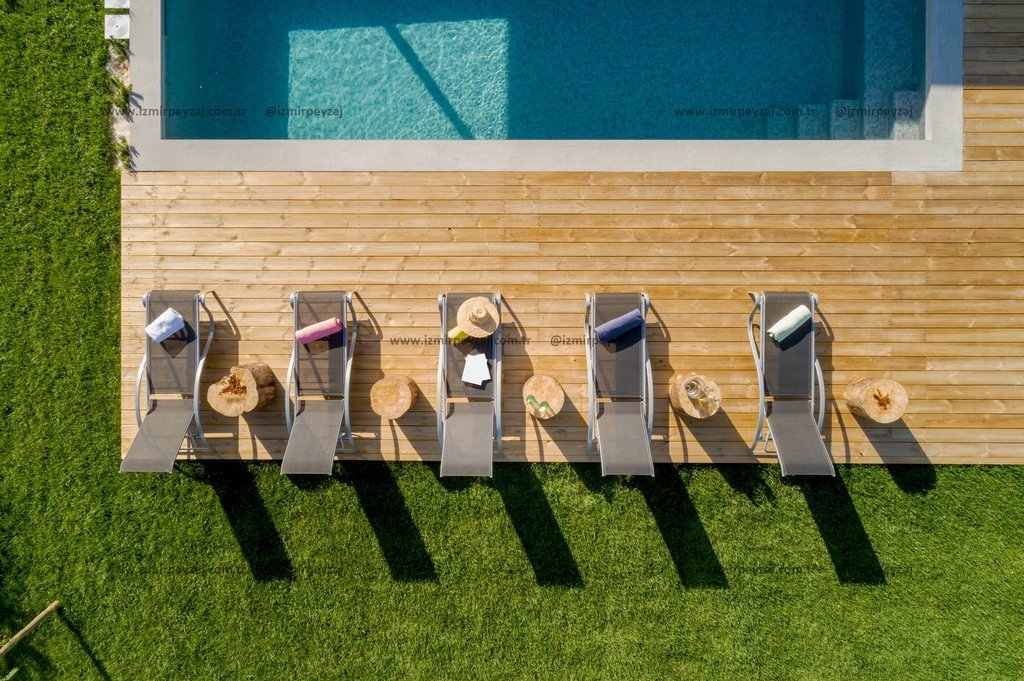 Havuz kenarında ahşap zemin üzerinde sıralanmış şezlonglar ve yeşil çim alanın kuşbakışı görüntüsü. Tatil ve dinlenme temalı bu görsel, huzurlu bir at