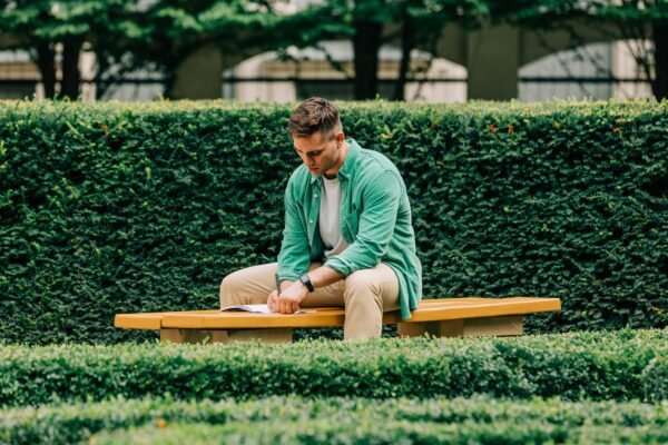 Stylish student guy in green shirt sits on bench in garden and write something